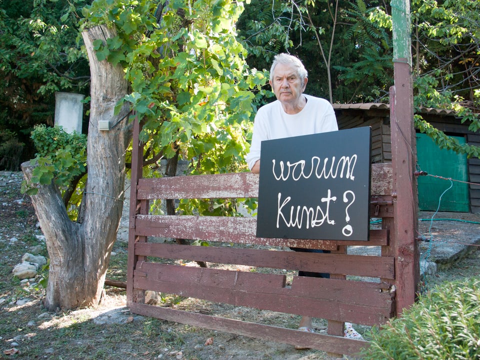 Ben Vautier lehnt sich an ein Gartentor. Er hat ein Schild in der Hand, auf dem «warum Kunst?» steht. 