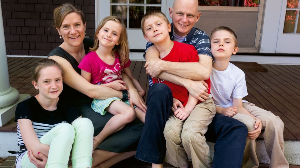 Eine Mann und eine Frau sitzen mit vier Kindern auf einer Veranda und lächeln.