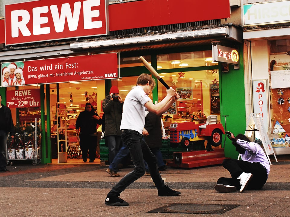 Vor einem Supermarkt bedroht ein Typ mit Baseballschläger einen Mann, der am Boden liegt. 