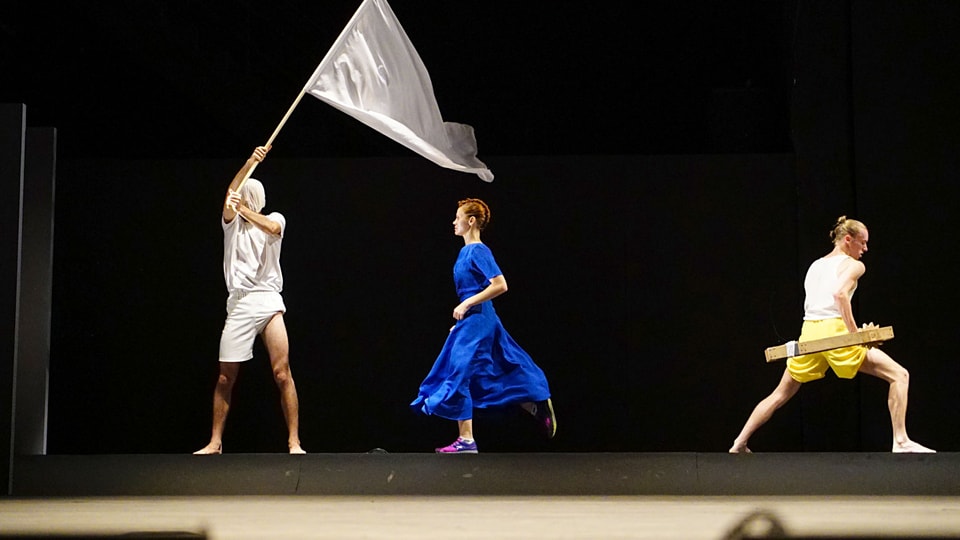 Drei Personen auf der Bühne. Eine mit einbandagiertem Kopf eine Fahne schwingend, eine Frau in einem leuchtend blauen Kleid und Turnschuhen, ein Mann trägt um die Hüften eine Holzkonstruktion.