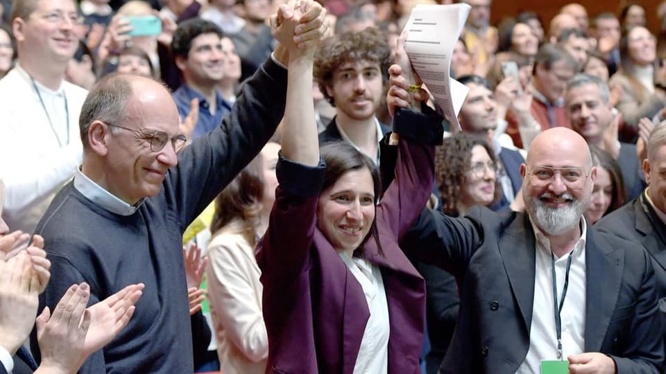Elly Schlein bei ihrer Wahl zur Parteichefin im vergangenen März. Zwei weisse Männer halten ihre Hände hoch.