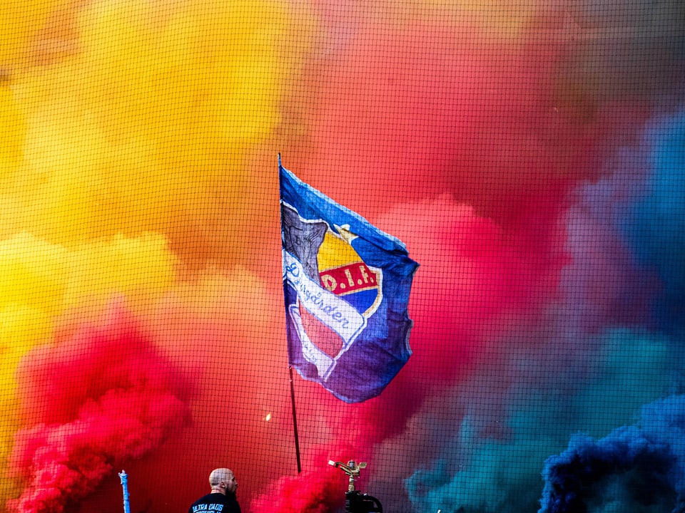 Djurgarden-Fahne ragt aus gelbem, rotem und blauem Rauch heraus.