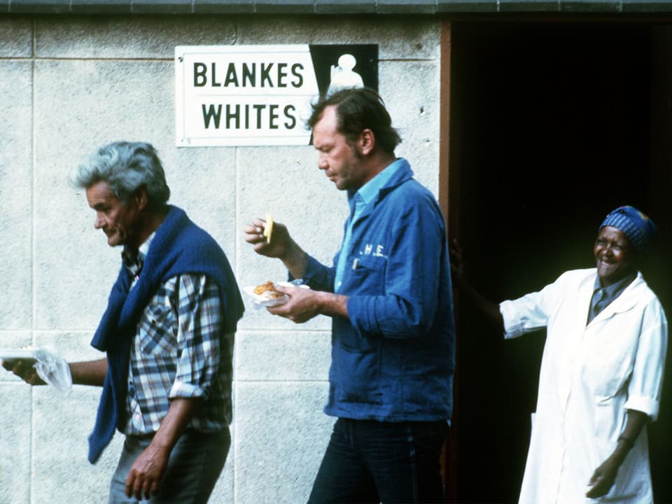 Zwei Weisse verlassen eine öffentliche Toilette mit dem Schild «Blankes/Whites», eine schwarze Toilettenfrau schaut ihnen nach.