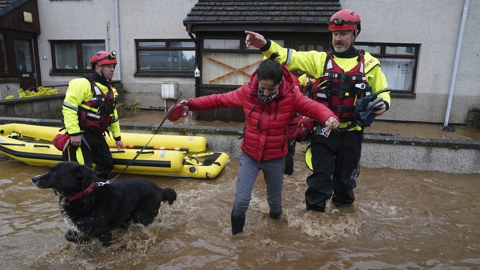 Frau watet mit ihrem Hund durchs Wasser. Sie wird von zwei Rettungskräften mit Gummiboot begleitet.
