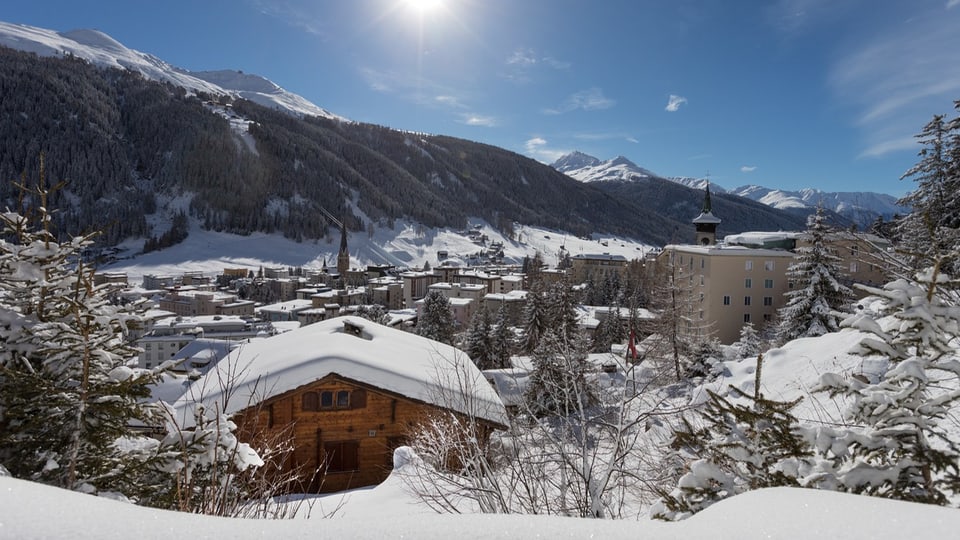 Panoramabild vom verschneiten Davos, vorne ein Holzchalet, hinten verschneite Hügel.