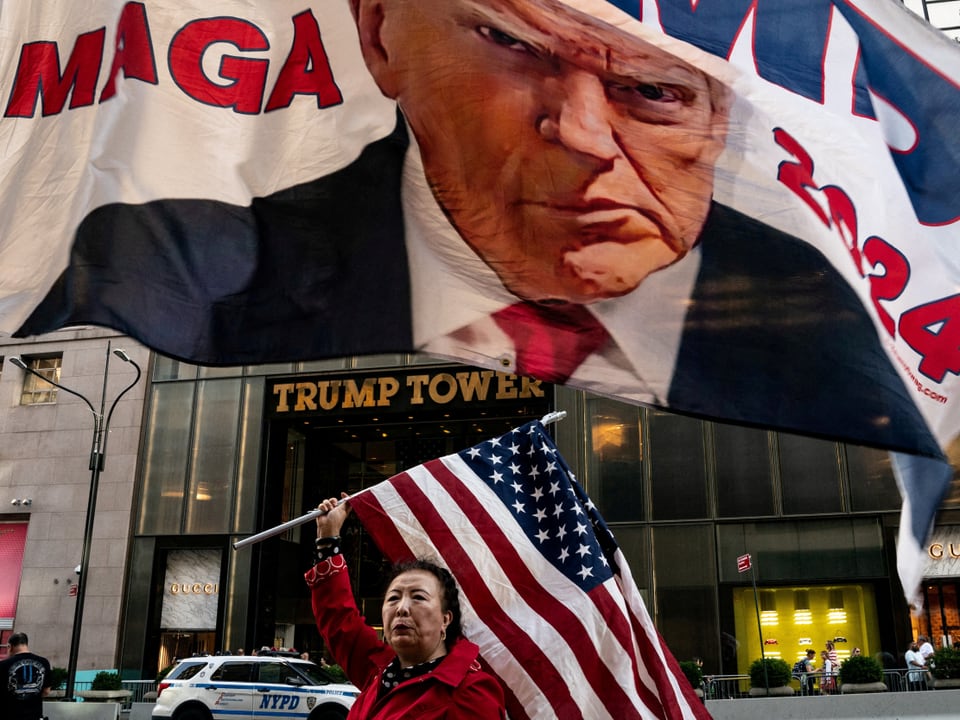 Eine Frau hält die amerikanische Flagge vor dem Trump Tower. Über ihr ist weht eine Wahlflagge mit Trumps Gesicht.