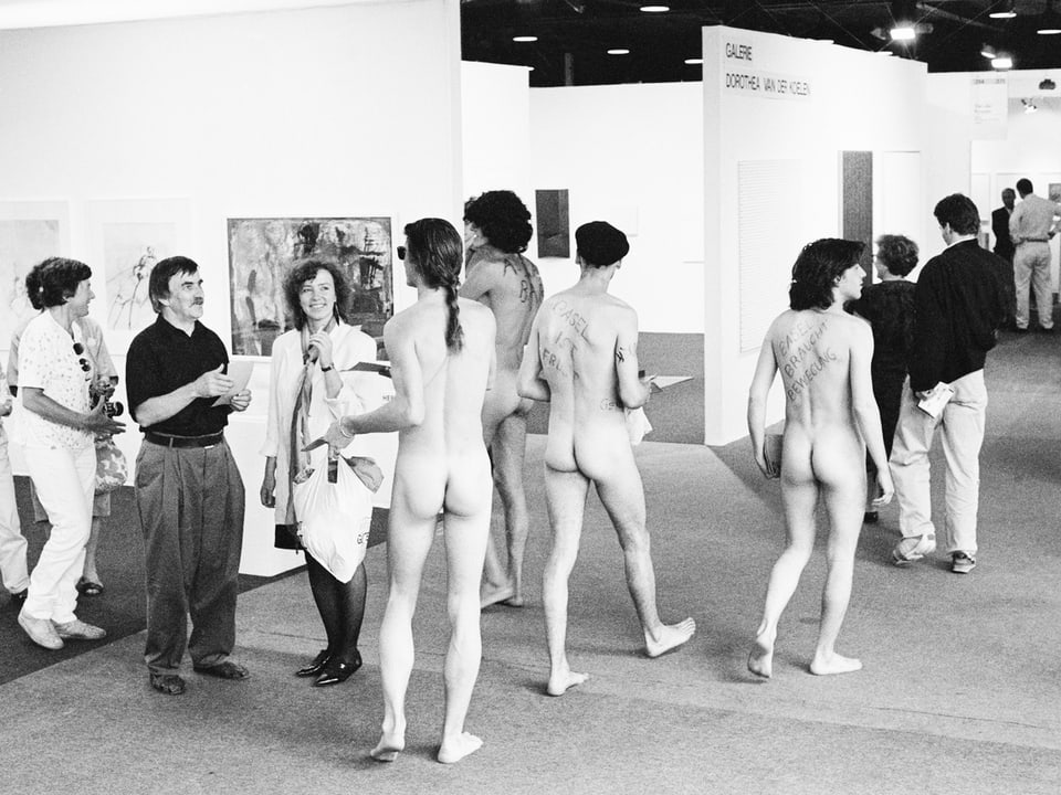 Eine Gruppe nackter Männer verteilt 1989 Flyer an lächelnde Besucher der «Art Basel» (Schwarzweiss).