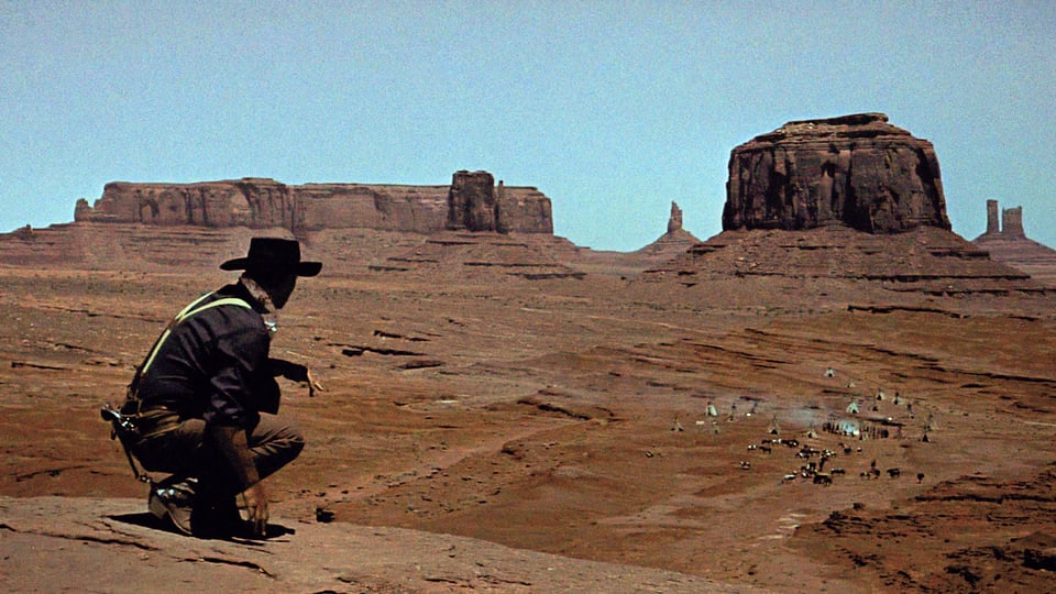 Filmsezene: Ein Mann schaut in einer Wüstenlandschaft auf eine weit entfernte Siedlung mit Zelten.