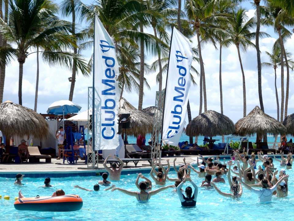 Wassergymnastik in einem Pool, im Hintergrund zwei Fahnen von Club Med, Palmen und schattenspendende rund Strohdächer