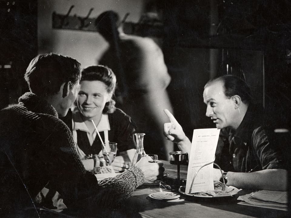 Franz Schnyder sitzt mit zwei Schauspielern (Einem Mann und einer Frau) am Tisch und gibt Regieanweisungen.