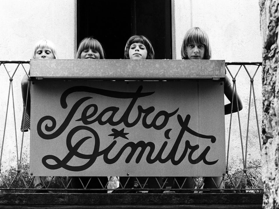 Schwarzweiss-Fotografie: Vier Kinder strecken ihre Köpfe hinter einem Schild hervor, auf dem «Teatro Dimitri» steht.