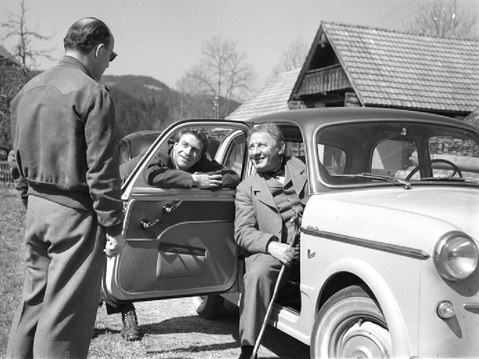 Franz Schnyder spricht mit zwei Schauspielern. Einer von ihnen sitz mit nach hinten geöffneter Beifahrertür im Auto, der andere lehnt sich nach vorne gegen ebendiese.