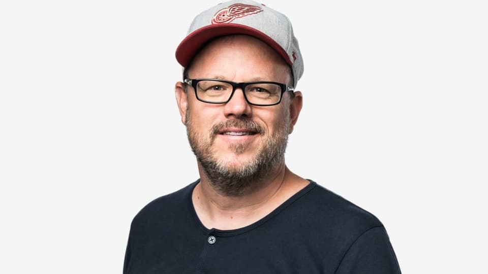 Phil Delisle lächelt in die Kamera, auf dem Kopf hat er ein Cap. Er trägt eine schwarze Brille und ein schwarzes Shirt.