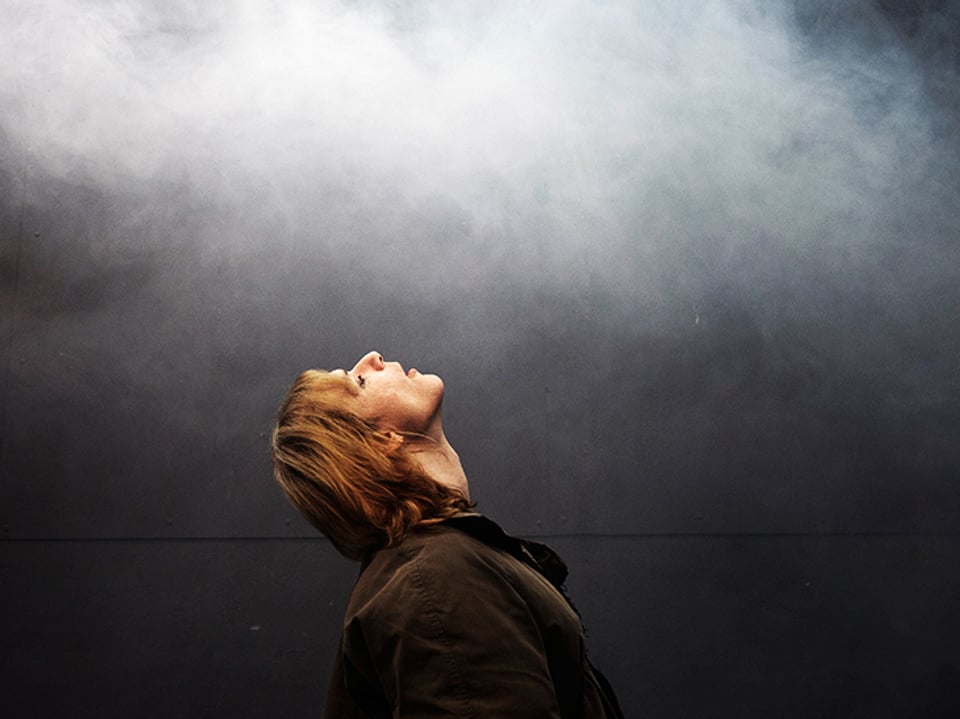 Eine Frau hält den Kopf im Nacken, das Gesicht gegen oben, der Mund ist liecht geöffent, oben hat es Nebel.