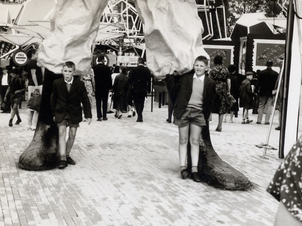 Schwarz-Weiss Fotografie mit zwei Buben, die neben den Füssen des Riesen Gulliver stehen.