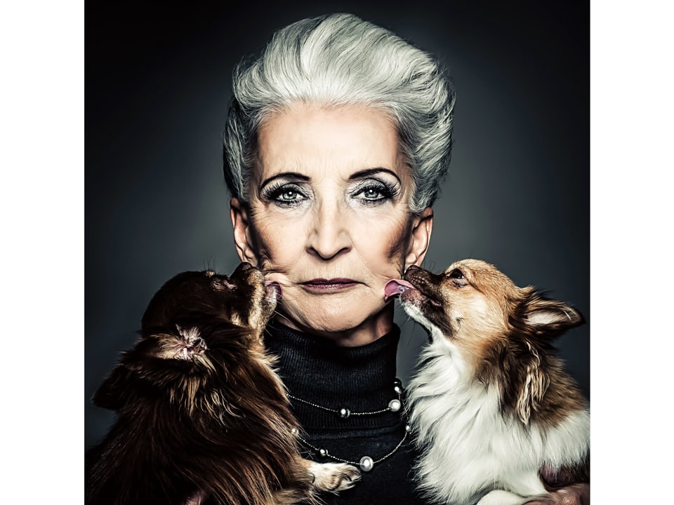Ältere geschminkte Frau, ihre Wangen werden von zwei Chihuahuas abgeleckt