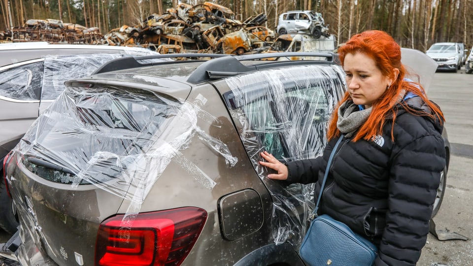 Eine Frau steht vor einem durch den Krieg in der Ukraine beschädigten Auto.