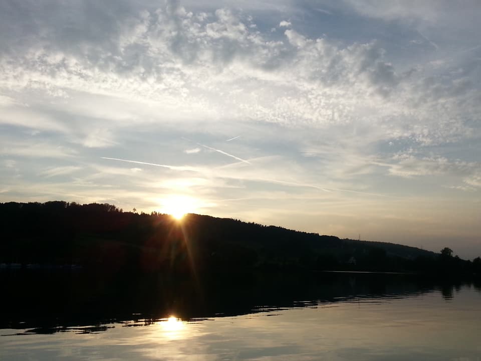 Spiegelblanker See im Vordergrund während hinter dem Zürichberg geht die Sonne unter. Am Himmel hat es harmlose hohe Wolkenfelder.