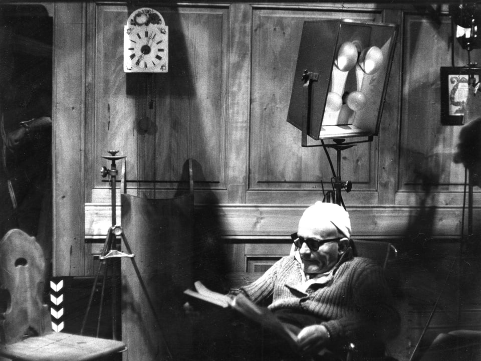 Franz Schnyder sitzt in einem Sessel und studiert das Drehbuch. Hinter ihm auf einem Stativ eine Filmlampe. Daneben ein weiteres Stativ und dahinter die Filmklappe an die Wand gelehnt.