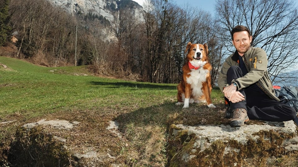 Nik Hartmann und Hündin Jabba sitzen auf einem grossen Stein in einer Berglandschaft.