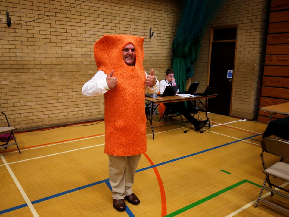 Mann in orangem Fischstäbchen-Kostüm in Turnhalle mit Wahl-Urne.