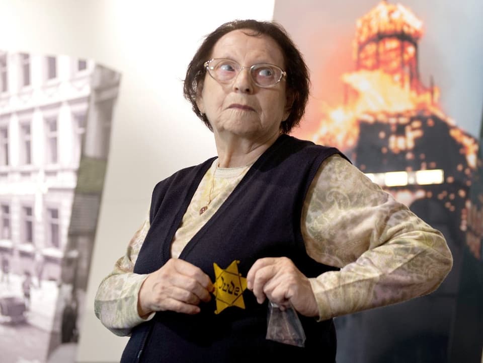 Eine Frau zeigt ihren Judenstern. Sie steht vor einem Bild mit brennendem Gebäude.
