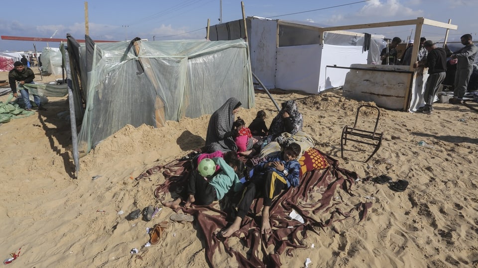 Zwei Frauen mit vier Kindern sitzen auf einer Decke im Sand, ohne Dach