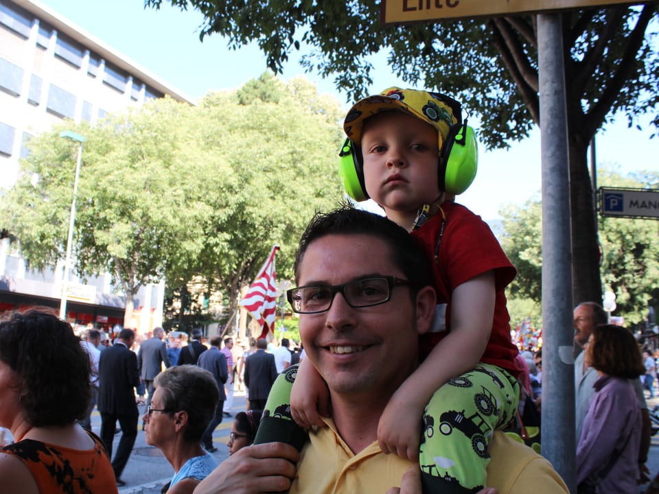 Mann mit Kind auf den Schultern, Kind hat Gehörschutz auf. Im Hintergrund Zuschauer des Festumzuges.
