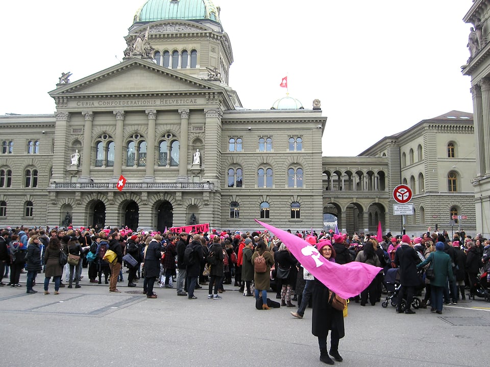 Im Vordergrund eine Frau, die eine rosa Fahne schwingt, im Hintergrund eine Menschenmenge vor dem Bundeshaus.