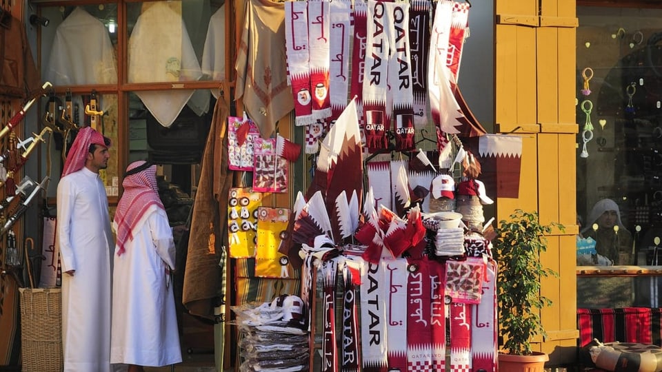 Rechts rot-weisse Schals mit der Aufschrift «Qatar», links Männer in weissem Gewand und mit Tuch bedeckten Köpfen.