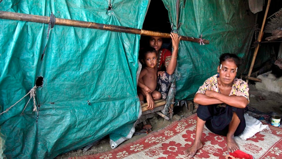 Zwei Flüchtlingskinder in einer Hütte aus Planer, davor sitzt eine Frau.