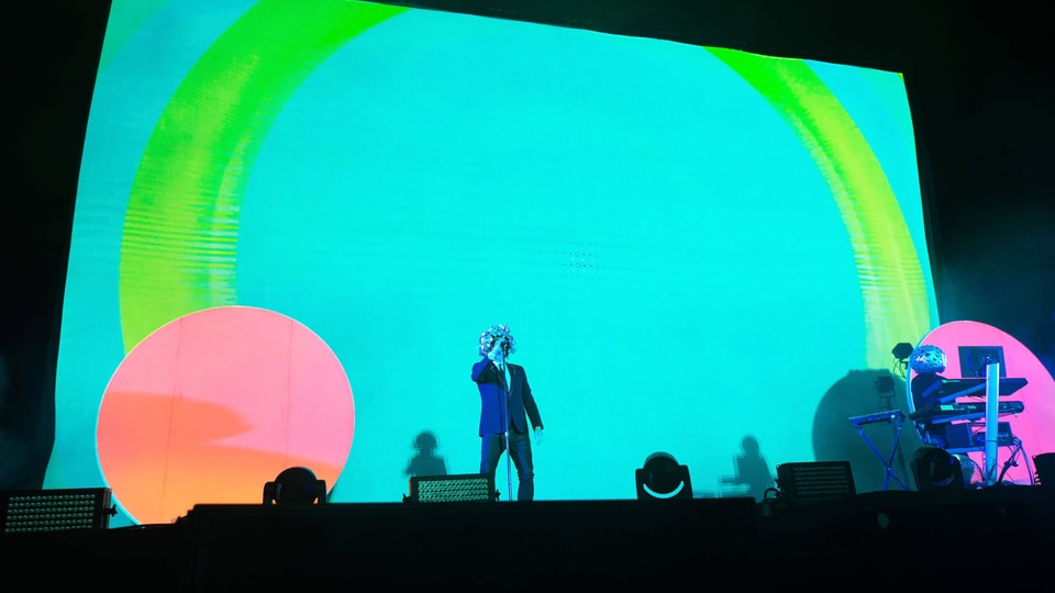 Die Pet Shop Boys auf einer schrillen Bühne: der Hintergrund leuchtet türkis, rosa und grün.