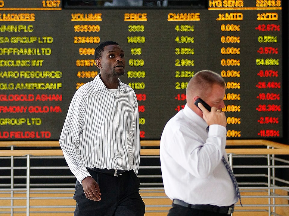 Ein Schwarzer und ein Weisser (Handy am Ohr) vor einer Anzeigetafel der Börse Johannesburg.