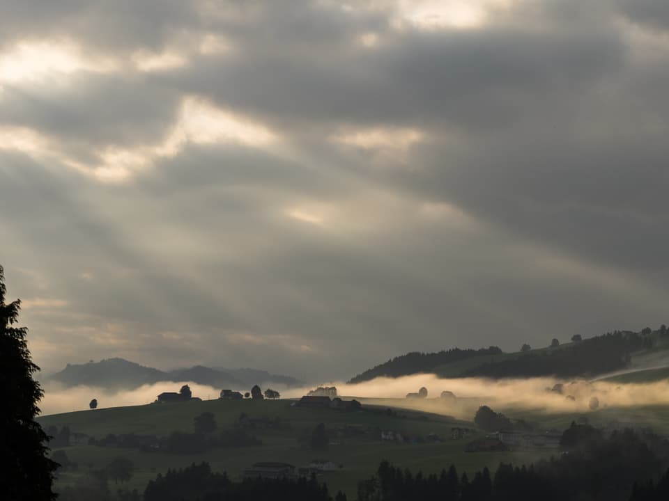 Die vielen grauen Wolken am Himmel lassen nur wenige Sonnenstrahlen durchschimmern, diese sind dafür als Lichstrahlen zu erkennen. Auf den Hügeln liegt zum Teil Nebel, welcher von der Sonne beschienen wird.