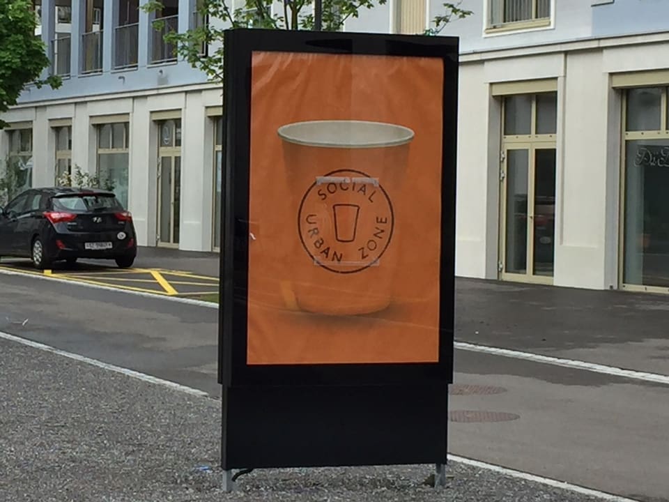 Ein viereckiges Schild steht auf einem Kiesplatz, darauf ist ein oranger Kaffeebecher zu sehen auf dem wiederum steht: «Social Urban Zone».