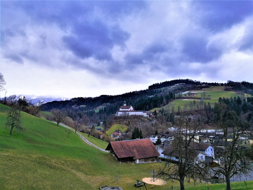 Hügelige Landschaft mit grünen Wiesen und ein paar Bäumen, im Tal Häuser und das Kloster.