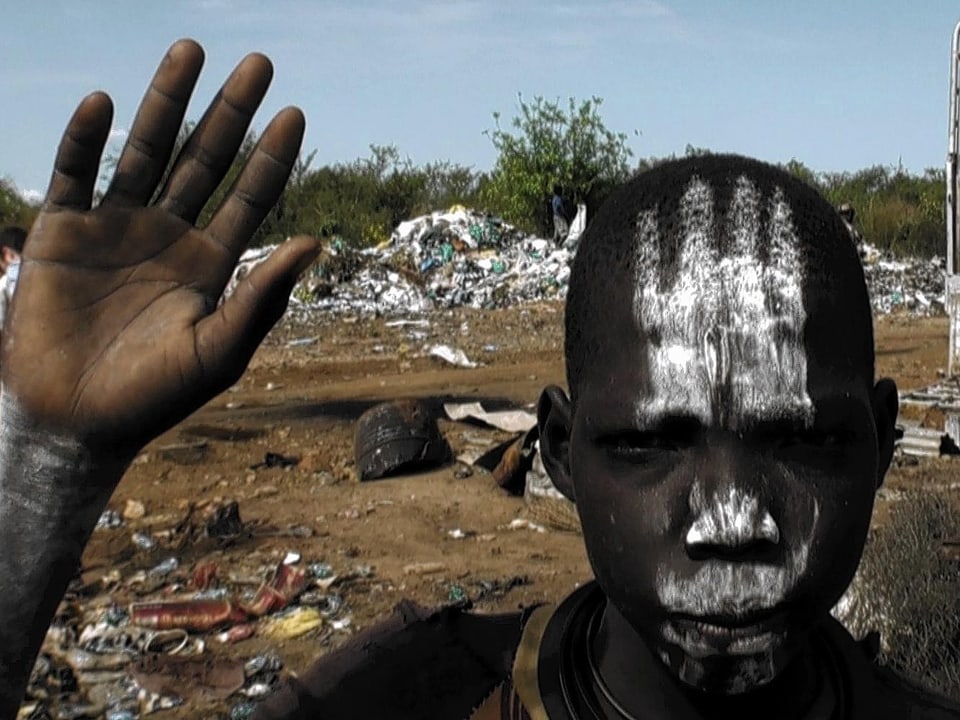 Ein sudanesischer Junge hebt seine Hand. Er blickt direkt in die Kamera und hat eine Gesichtsbemalung.
