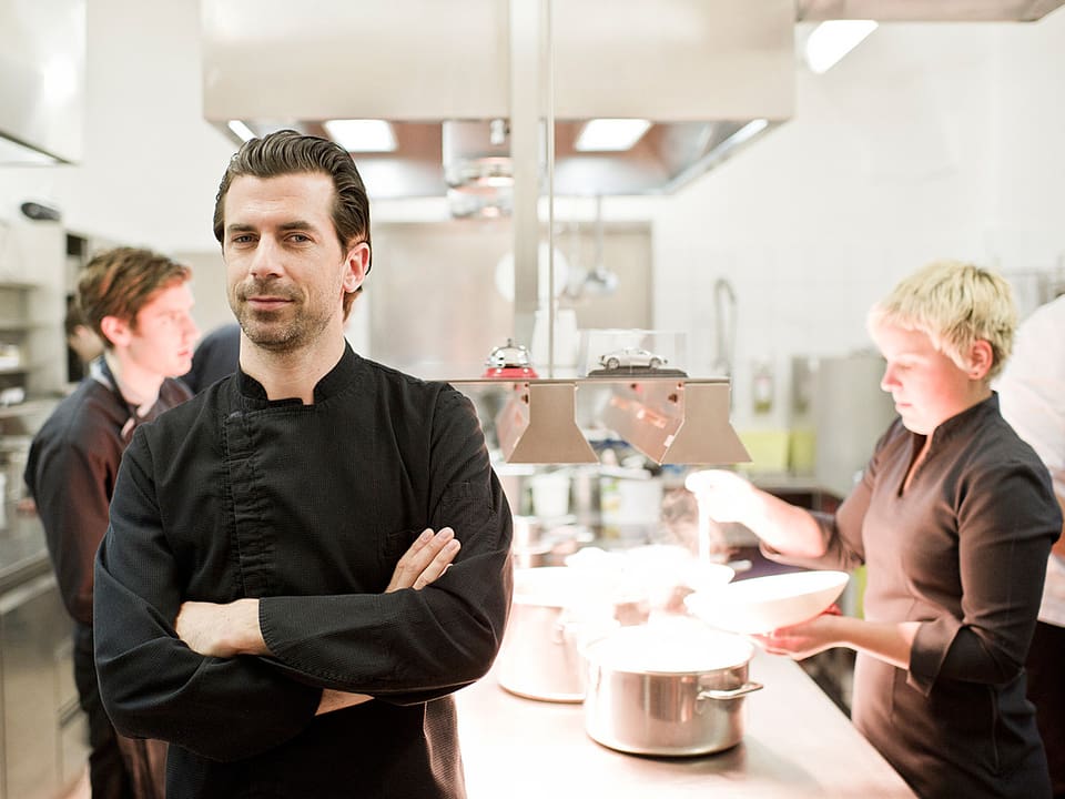 Porträt von Andreas Caminada, im Hintergrund sind eine Köchin und ein Koch am Werk.