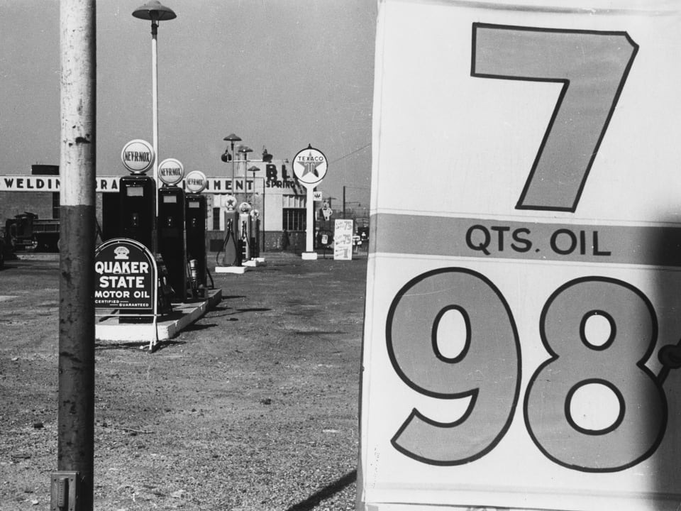 Auf einer schwarz-weissen Fotografie ist eine verlassene Tankstelle zu sehen: Zapfsäulen, rechts ein Schild mit den Ziffern sieben und 98.