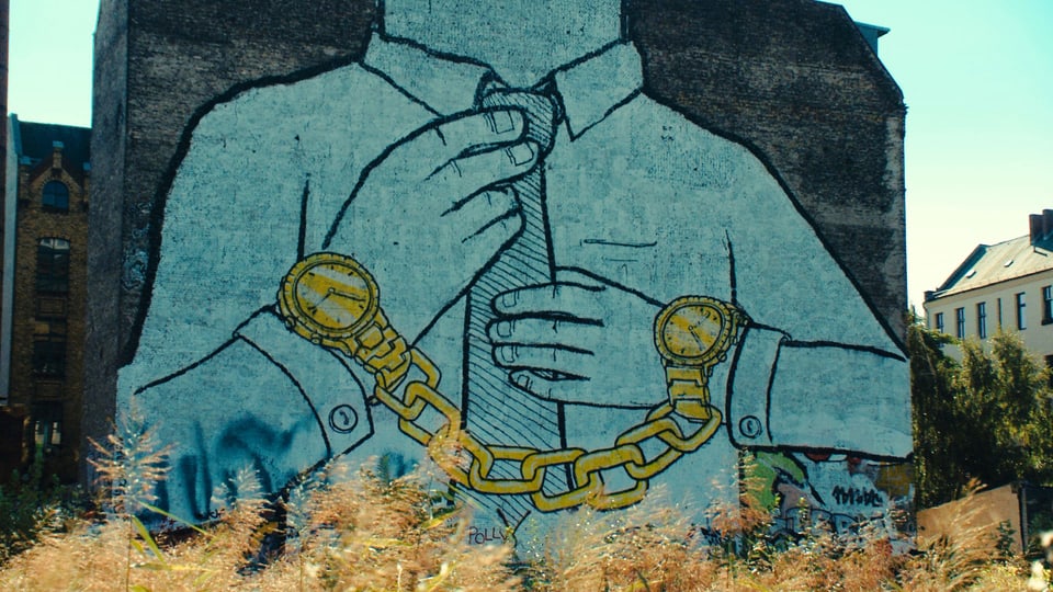 Ein an eine Hauswand gemaltes Bild zeigt einen Mann mit Krawatte, der an beiden Armen eine Uhr trägt; dabei sind die beiden Uhren mit einer schweren Kette verbunden.