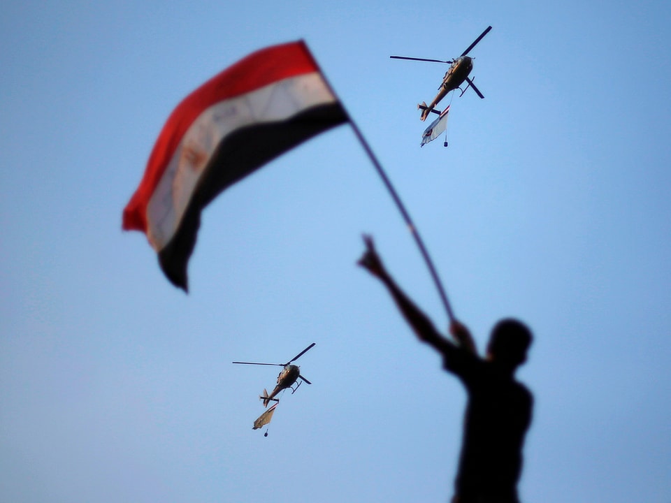 Zwei Helikopter mit Fahnen in der Luft. Im Vordergrund schwenkt ein junger Mann eine ägyptische Fahne.
