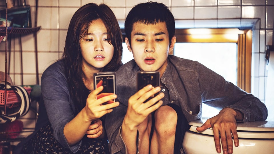 Filmszene: Zwei junger Mann und eine junge Frau sitzen neben einer Toilettenschüssel und schauen aufs Smartphone.