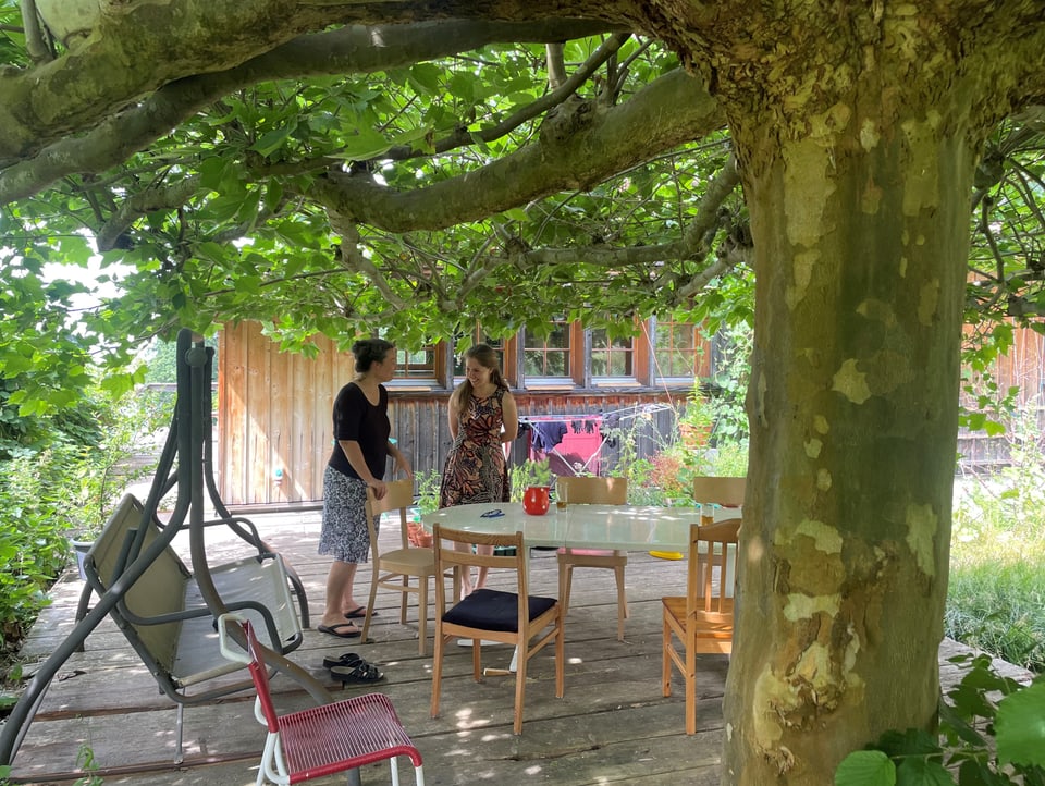 Im Schatten eines grossen Baumes steht ein Tisch mit Stühlen und eine Schaukelbank. Zwei Frauen unterhalten sich.
