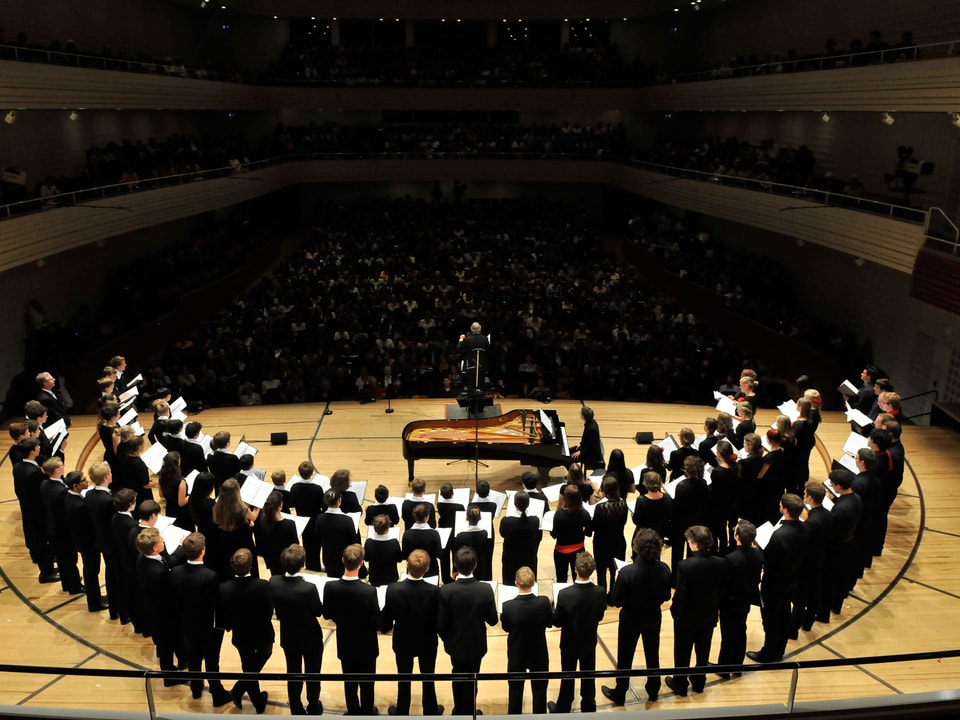 Ein Chor steht in einem Halbkreis auf der Bühne auf dem dunklen Saal.