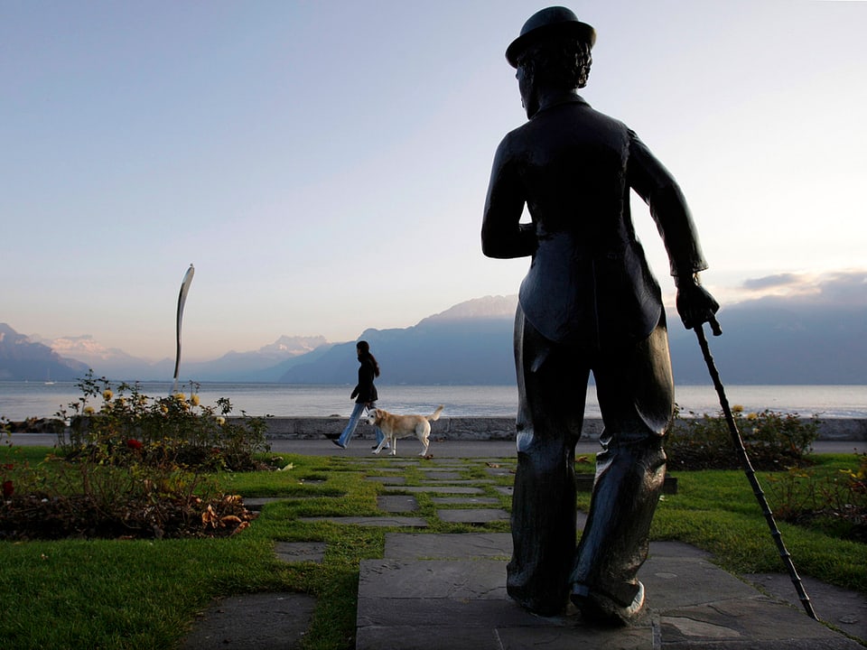 Statue von Charlie Chaplin in Vevey. Im Hintergrund sind der Genfer See und Berge erkennbar.