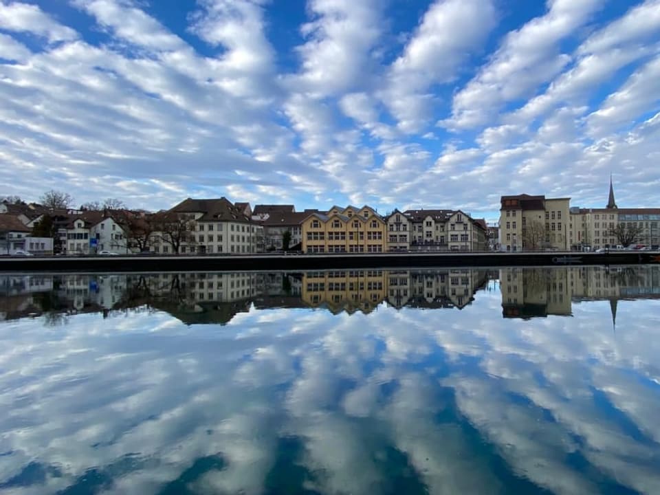 Blick über einen Fluss auf Stadt. Der Himmel blau mit weissen Streifen, die sich im Wasser spiegeln. 