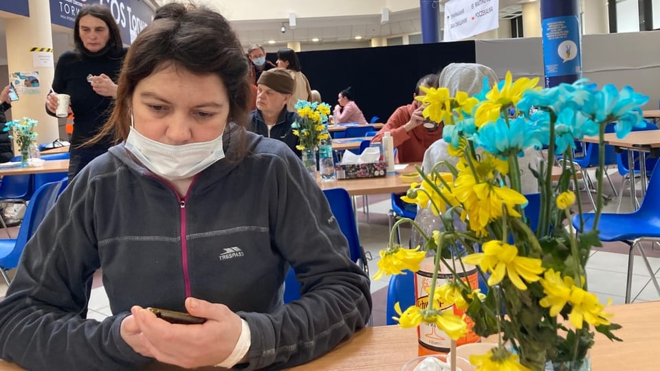 Eine Frau sitzt an einem Tisch, Handy in der Hand, gelbe und rote Blumen in einer Vase daneben.