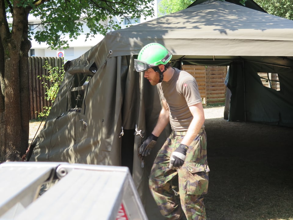 Mann in Militär-Kleidern mit Grünem Helm und Schutzbrille tritt aus Zelt