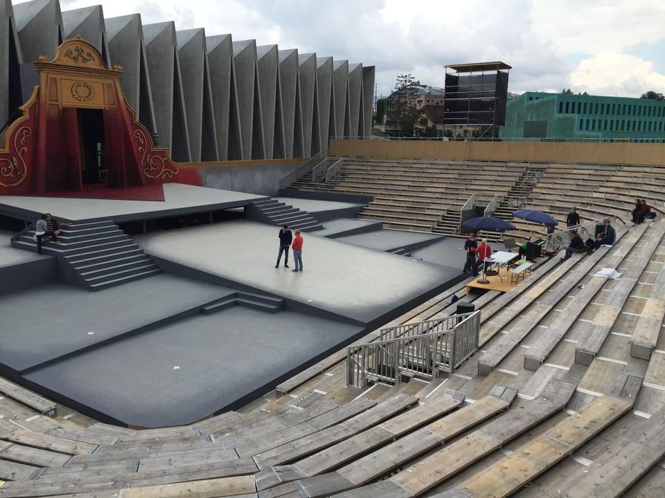 Arena mit Holzpritschen und Bühne, dahinter ein modernen Beton-Bau
