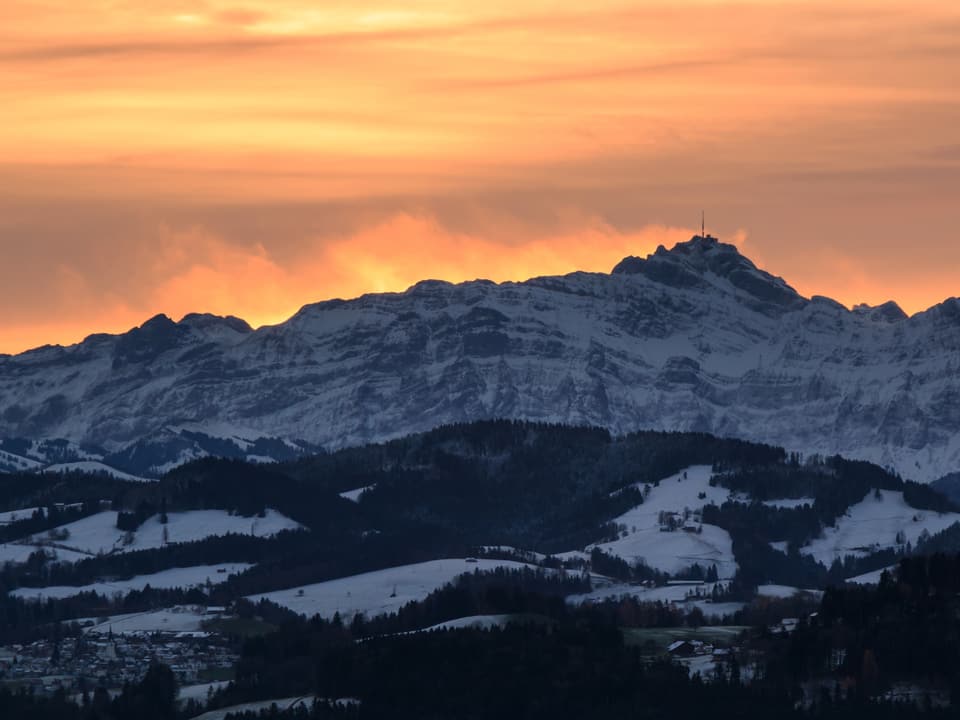 Eingeschneite Bergkette mit Windfahnen an den Gipfeln. Diese werden von der Sonne orange beleuchtet. Der Himmel ist orange gefärbt.